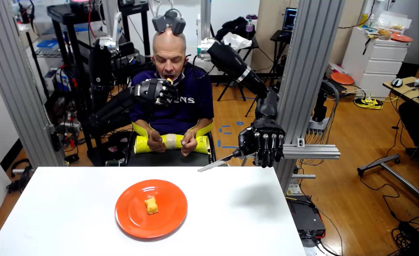 an zit op stoel in technische opstelling en krijgt te eten doordat twee robotarmen het eten naar zijn mond brengen.